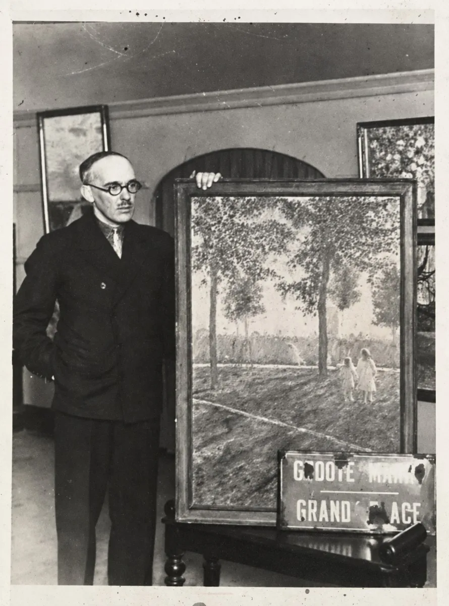 Black and white photograph of Gerard Ceunis, in glasses and a dark suit, standing beside one of his framed oil paintings of a tree-lined path with two small figures, with a Groote Markt Grand Place street sign propped in front
