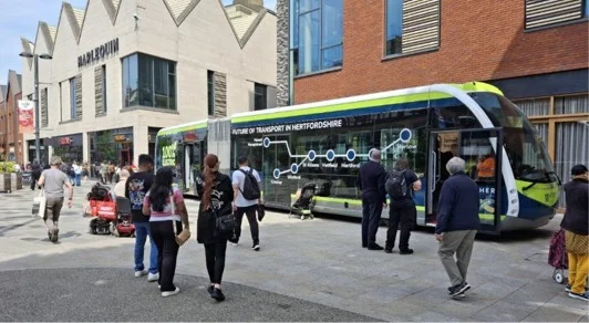 Computer-generated image of a HERT trackless tram in Future of Transport in Hertfordshire livery at a stop in Watford town centre, with people walking past