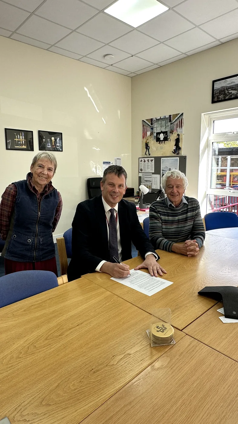 Headteacher Mark Nicholls signs the pledge at a meeting table, with Councillors Wren and Rowlands beside him.
