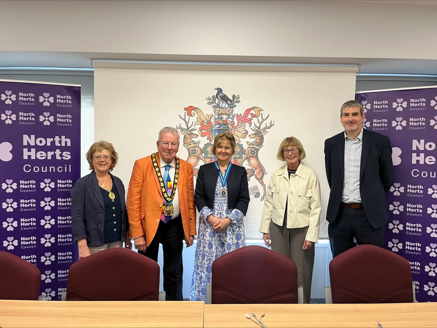 Five people standing in the North Herts Council chamber in front of the council's coat of arms and two purple North Herts Council banners
