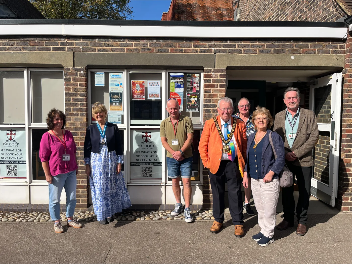 Seven people standing outside the brick front of Baldock Community Hub, its windows covered in posters for a quiz night and a trivia night