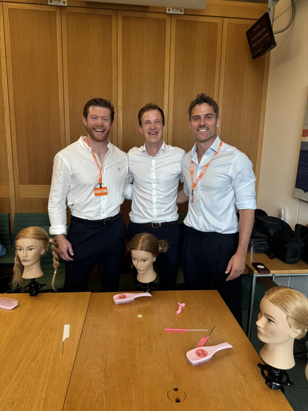 Three men in white shirts standing behind a table set with practice heads, pink hairbrushes and combs in a Parliament meeting room, two of them wearing orange visitor lanyards