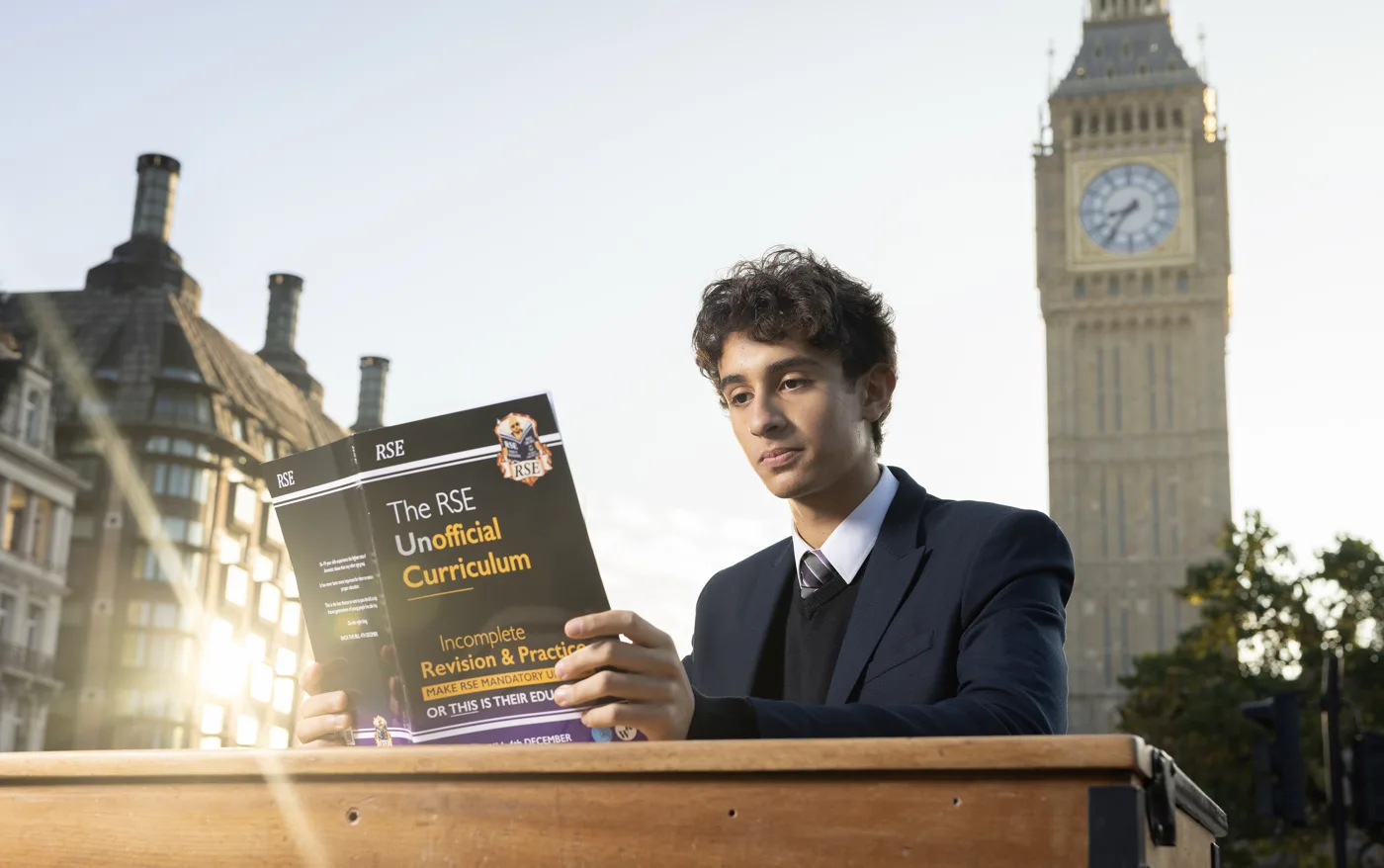 A young man in a school blazer reads a copy of the RSE Unofficial Curriculum at a wooden school desk in Parliament Square, with the Elizabeth Tower behind