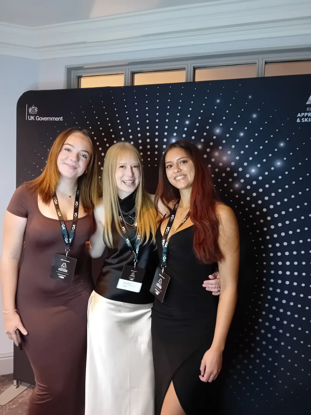 Three young women in evening dress and awards lanyards smiling in front of a Regional Apprenticeship and Skills Awards backdrop