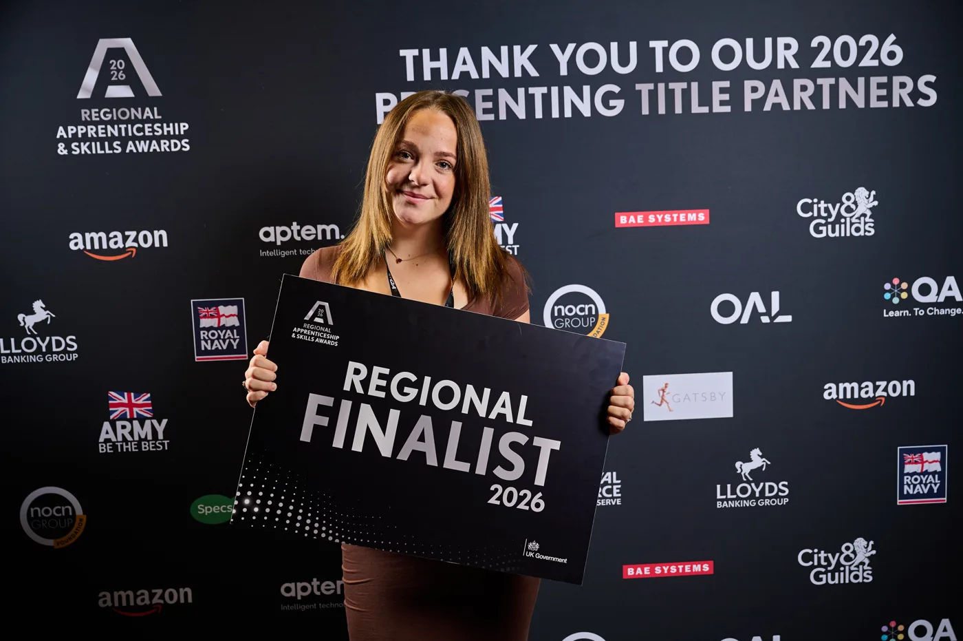 Lilly Ash holding a Regional Finalist 2026 sign in front of a sponsors' backdrop